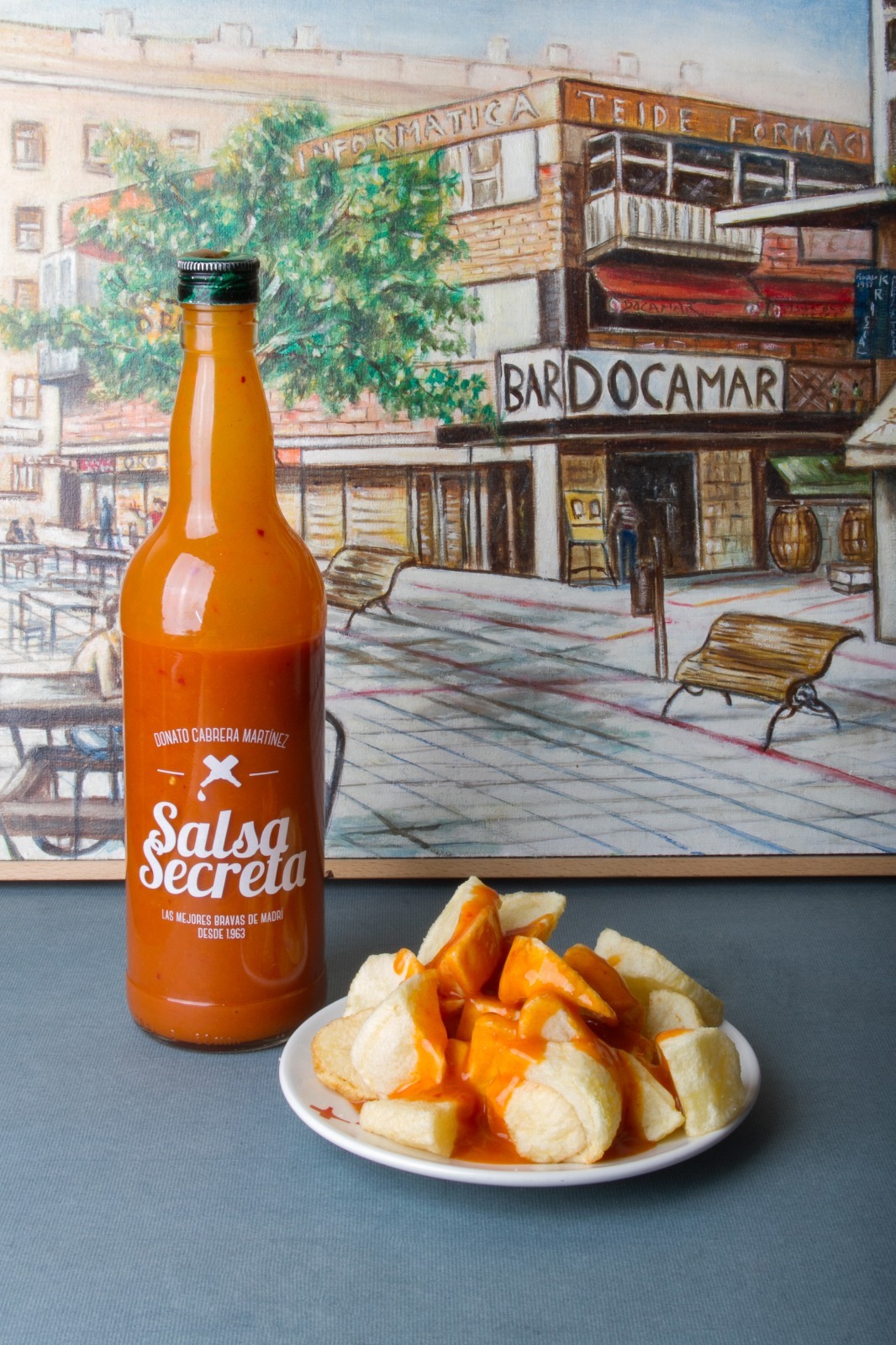 Botella de Salsa Secreta Docamar junto a un plato de bravas, ante el mural pintado de la fachada