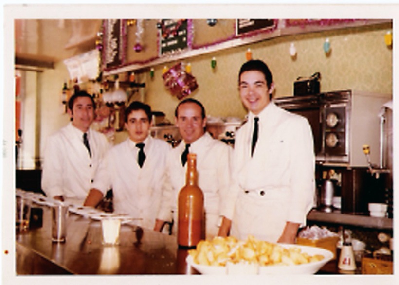 Cuatro camareros vestidos de blanco tras la barra, con una botella de whisky DYC y un plato de bravas